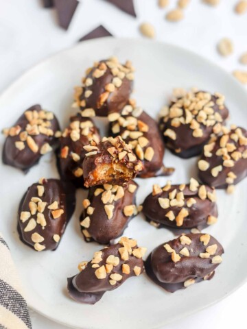 Snickers dates coated in dark chocolate and topped with granulated peanuts, served on a white plate.