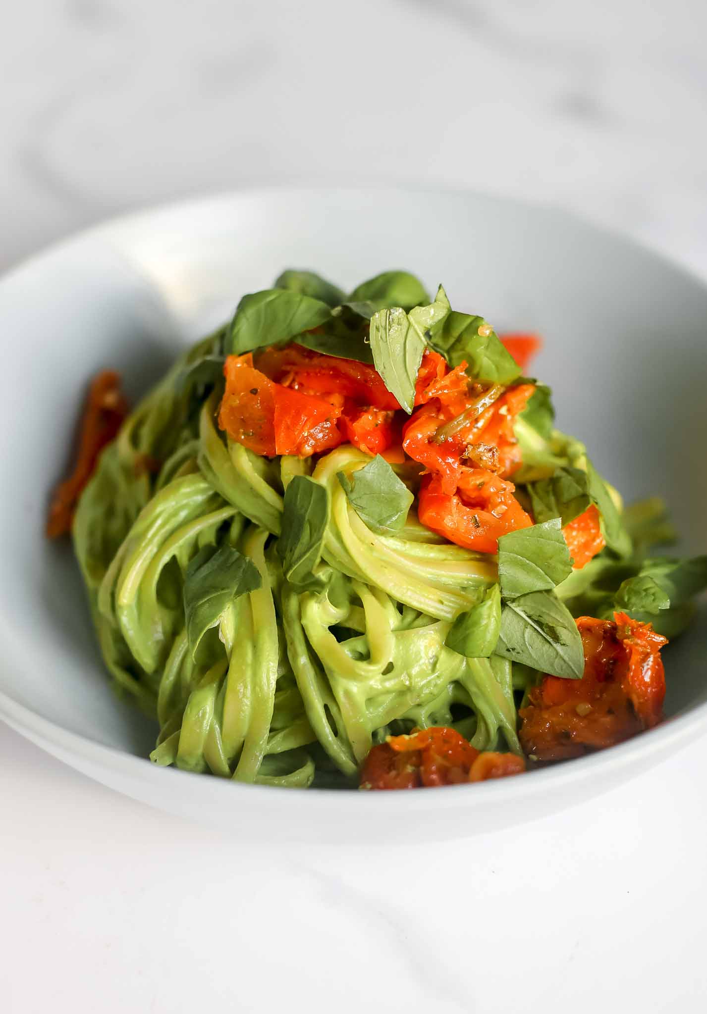 Bowl of avocado pasta on marble benchtop topped with sundried tomatoes and basil leaves.